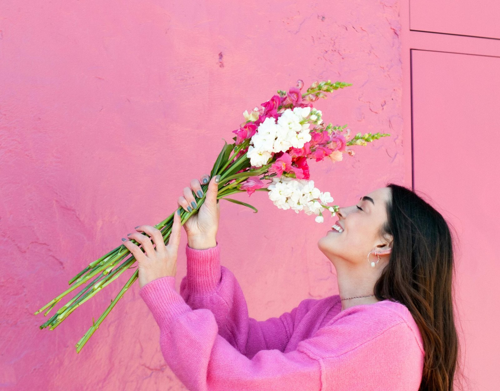 Side view of a brunette woman in a pink sweater enjoying a bouquet of gladiolus against a vibrant pink wall.